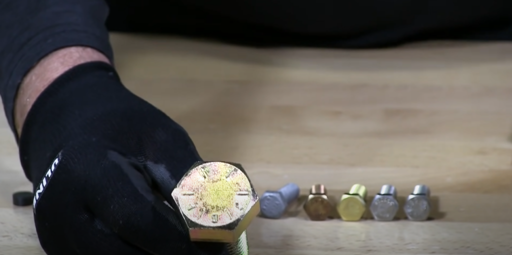 Assorted bolts on a table, with a gloved hand holding one bolt up close to the camera