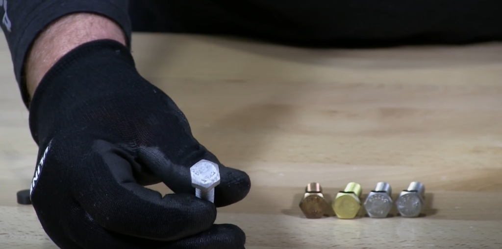 Assorted bolts on a table, with a gloved hand holding one bolt up close to the camera